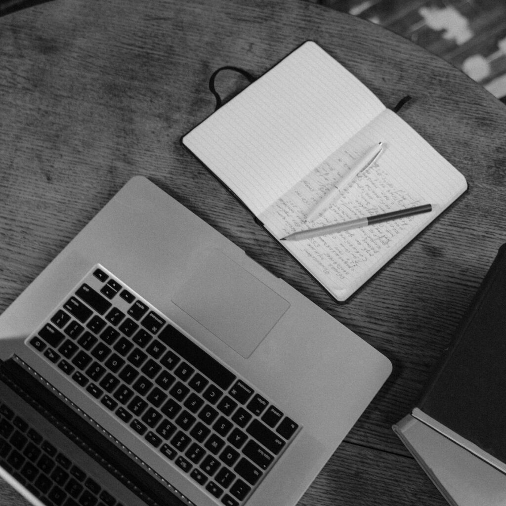 Black and white top view of a workspace with a laptop, notebooks, and pens on a wooden table.